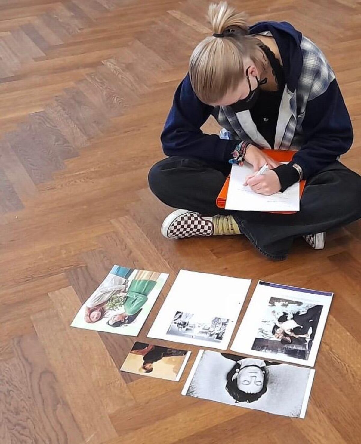 Eine Schülerin sitzt am Boden, vor ihr liegen unterschiedliche Bilder. Sie schreibt einen Text.