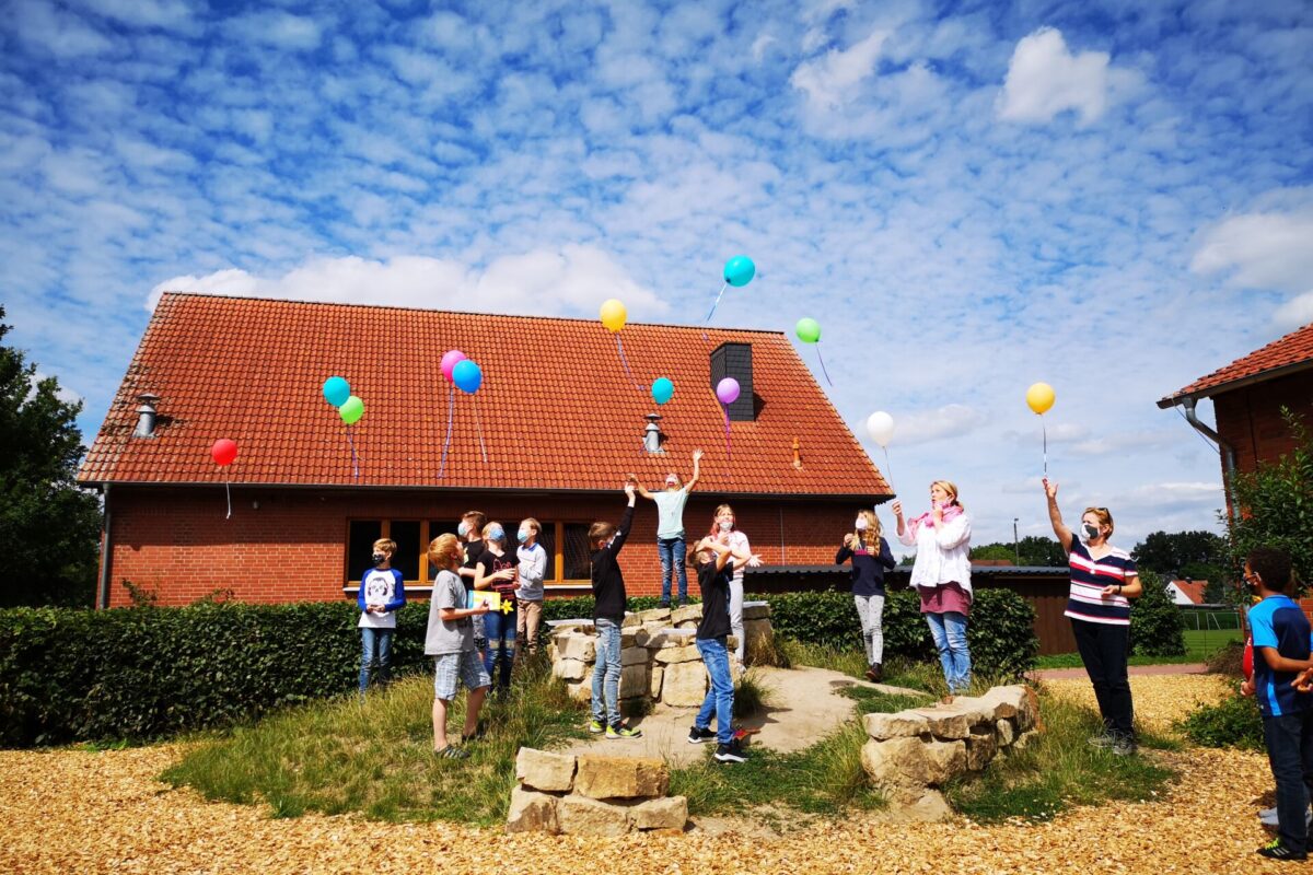 Kinder und Lehrerinnen stehen auf einem Hügel vor dem Schulgebäude und lassen Luftballons steigen